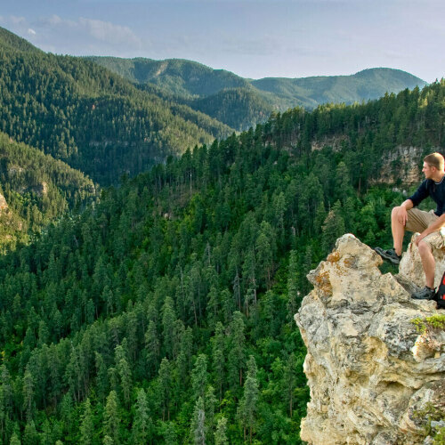 Hiker sitting on top of rim rock ledge overlooking pine tree valley 