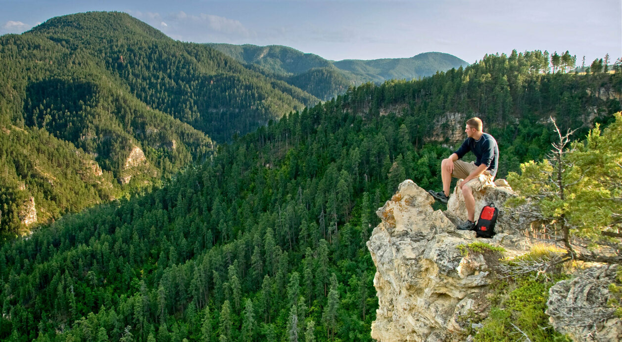 Hiker sitting on top of rim rock ledge overlooking pine tree valley 
