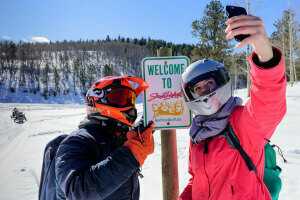 Two Black Hills Snowmobilers posing for a selfie in front of a welcome to South Dakota sign